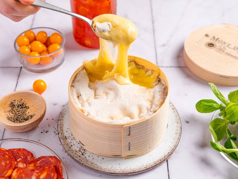 Dampet ostesmørbrød med smeltet ost som trekkes opp fra en rund bambusboks, omgitt av små skåler med tomater, krydder og urter på et kjøkkenbord.