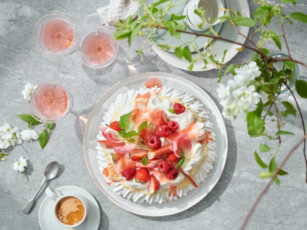 En tallerken med en dessert med jordbær, krem og frukt, omgitt av tre glass med rosa drikke og grønne planter.