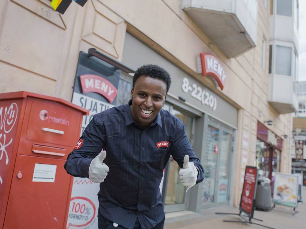 En person står foran en butikk og gir tommel opp, iført mørk skjorte og hvite hansker, med en rød postkasse i bakgrunnen.