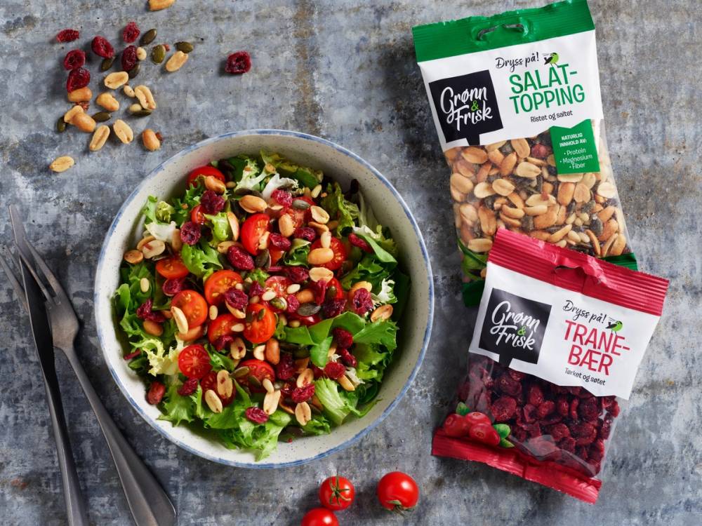 En frisk salat i en hvit bolle med ulike grønnsaker og nøtter, omgitt av to poser med salat topping og tranebær, på en grå steinplate.