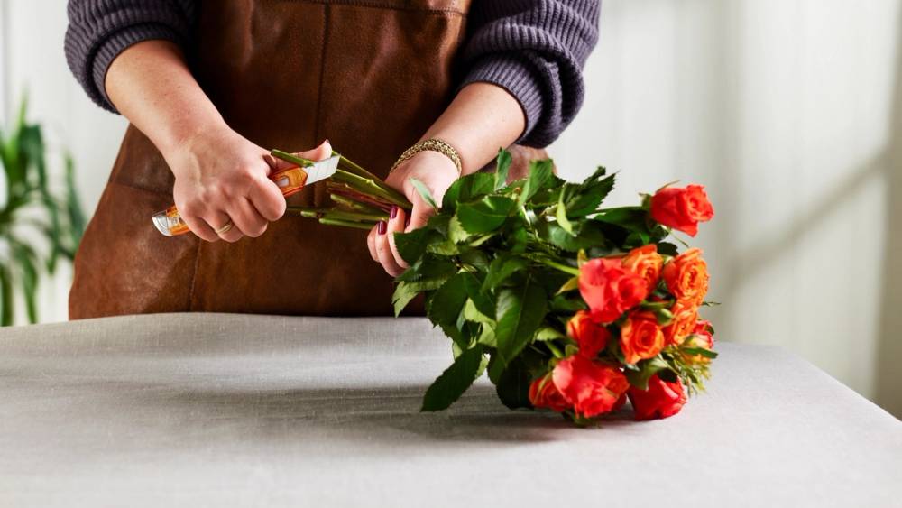 Person som klipper stilkene på en bukett med røde og oransje blomster på et bord.