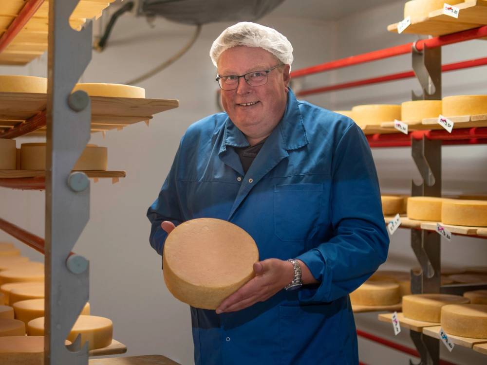 En person i blå arbeidsjakke ser på en stor ost i et rom med ostestativer og osteskiver på hyller.