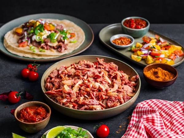 En tallerken med en ferdiglaget taco med kjøtt, grønnsaker og ost, omgitt av ulike små skåler med krydder og tilbehør, på et mørkt bord med noen cherrytomater og en rød og hvit rutete serviett.