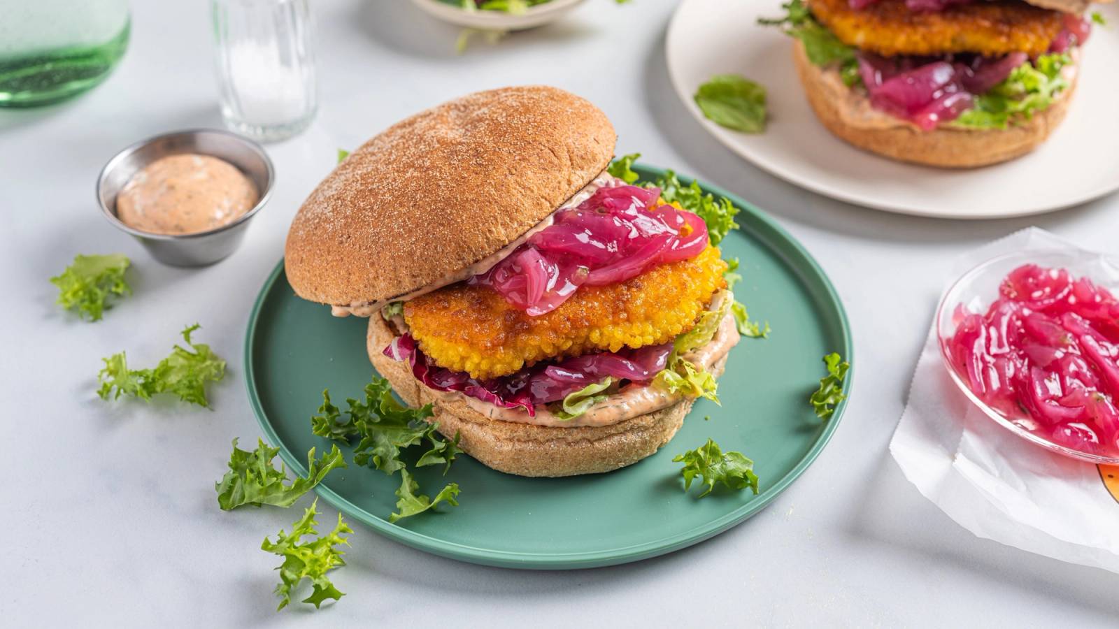 Sprø fiskeburger med syltet rødløk på en grønn tallerken, omgitt av små skåler med saus og grønnsaker.