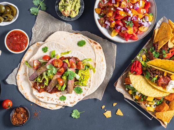 Tacos med kjøtt, salat og tomater, omgitt av salsa, guacamole og nachos på en mørk bakgrunn