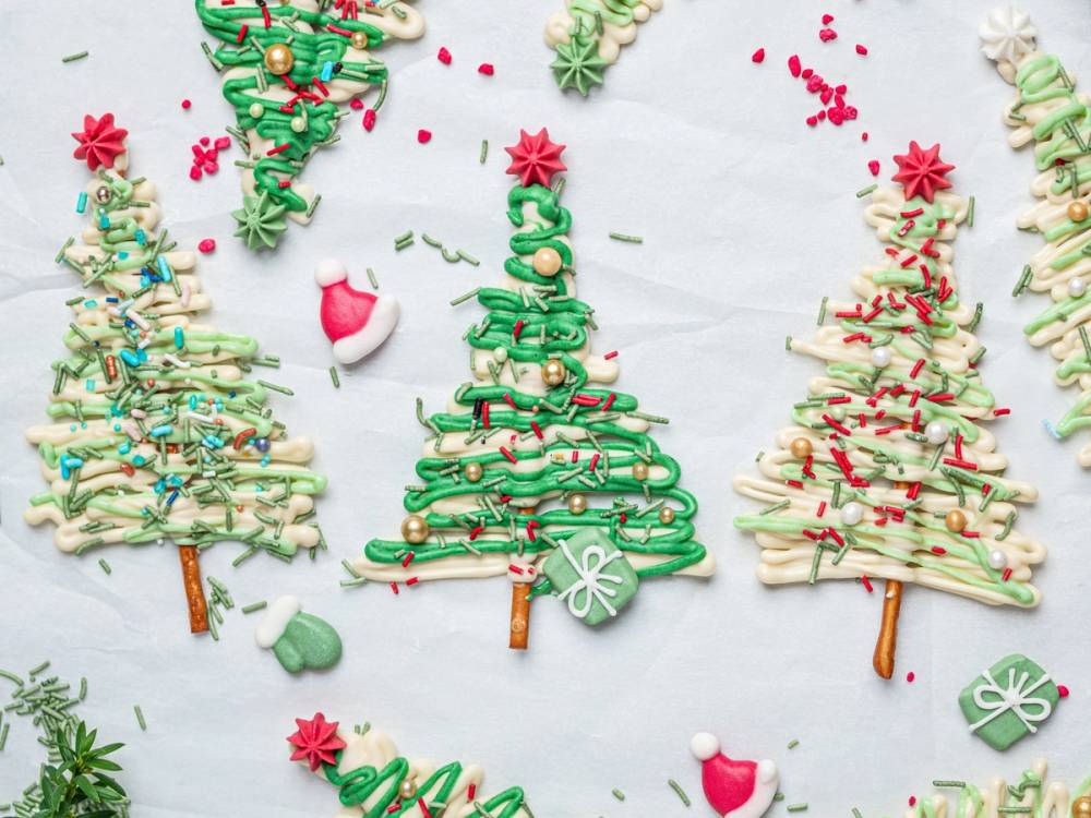 Små juletrær laget av sjokolade og pyntet med ulike fargerike striper og dekorasjoner, plassert på et hvitt underlag.