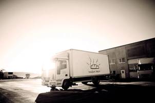 En hvit lastebil med en logo av et brød kjører forbi en industribygning i solnedgang, på Sandnes-bakeris område.