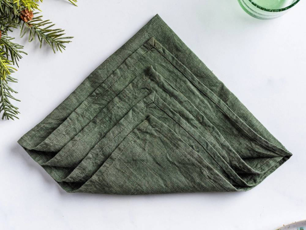 En grønn serviett brettet til et juletre, lagt på en hvit overflate med noen grønne grener i hjørnet.
