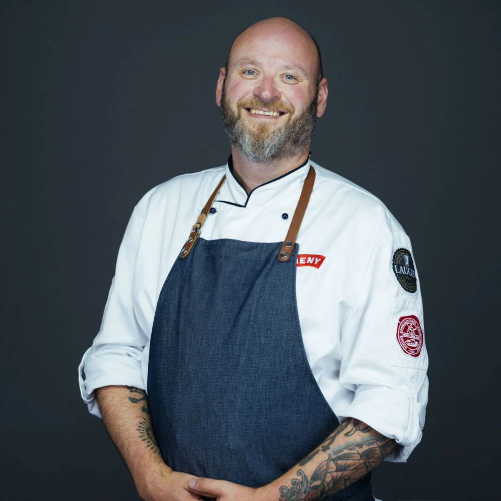 En person i kokkeuniform med en blå forkle og tatoveringer på armene, står mot en mørk bakgrunn.