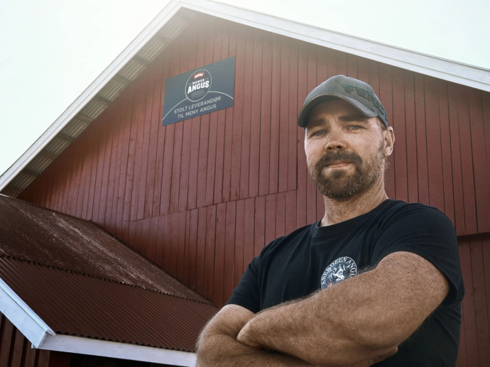 En person står foran en rød bygning med et skilt som sier 'Hovin Angus', og personen har armene krysset, iført svart t-skjorte og caps.