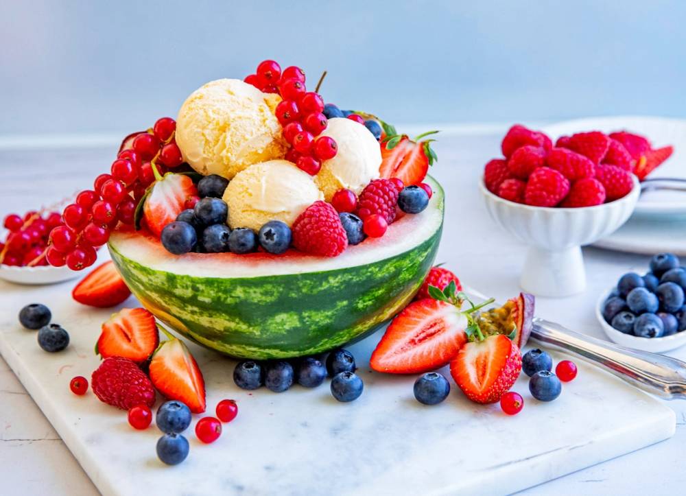 Vannmelonkake fylt med vaniljeis og toppet med ulike bær som jordbær, blåbær, bringebær og rips, med flere bær rundt på et hvitt bord.