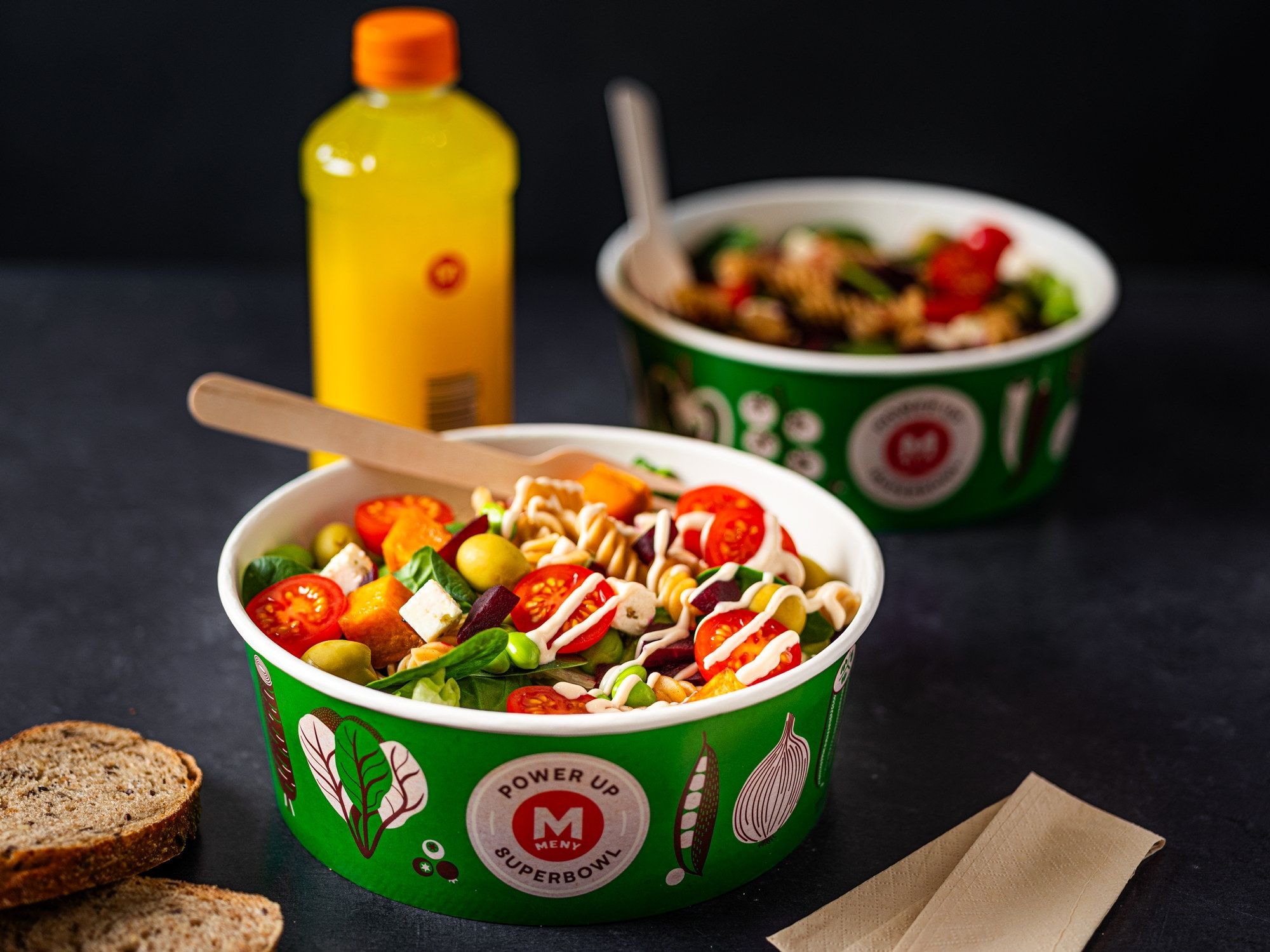 To bowls med salat med cherrytomater, agurk og mais, med en skje i hver, på et mørkt bord med brødskiver og en gul flaske i bakgrunnen.