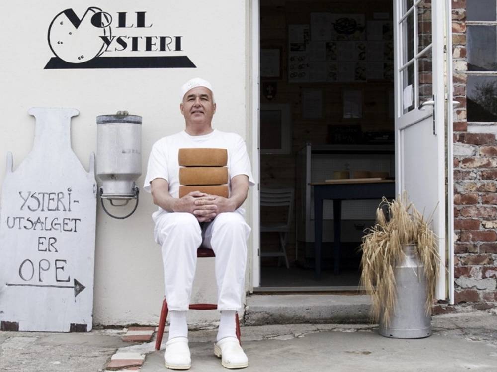 En person i hvit klær som sitter foran en ysteributikk med en skilt som sier at ysteriet er åpent, og det står en stor metallbeholder og en kornhaug ved inngangen.