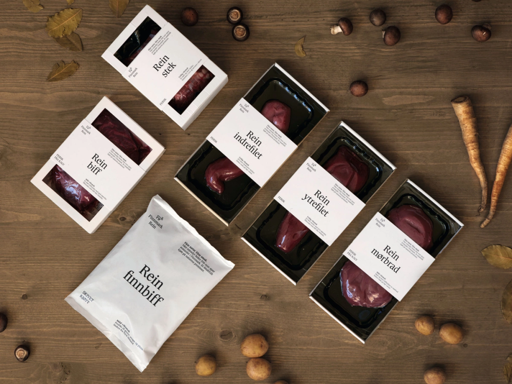 Pakker med reinsdyrkjøtt og en pose med reinsdyrkjøtt fra Finnmark, omgitt av nøtter og en trepinne på en treflate.