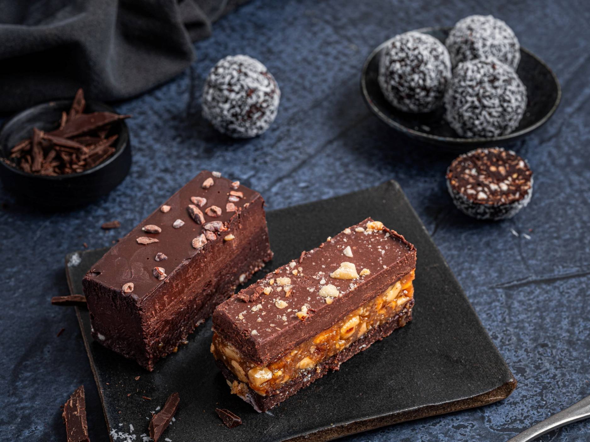 To kakerstykker med sjokolade og nøtter på en svart tallerken, omgitt av små kuler med kokos og en skål med sjokoladekuler, på en mørk overflate.