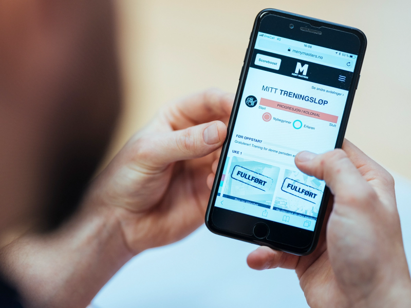 Person holder en smarttelefon som viser en nettside med overskriften 'MITT TRENINGSLOP' og ulike menypunkter, på en lys bakgrunn.
