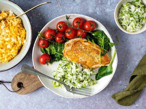 En tallerken med kokt ris, cherrytomater, en skive av stekt fisk, og grønne blader, med en liten skål med rømme eller saus ved siden av.