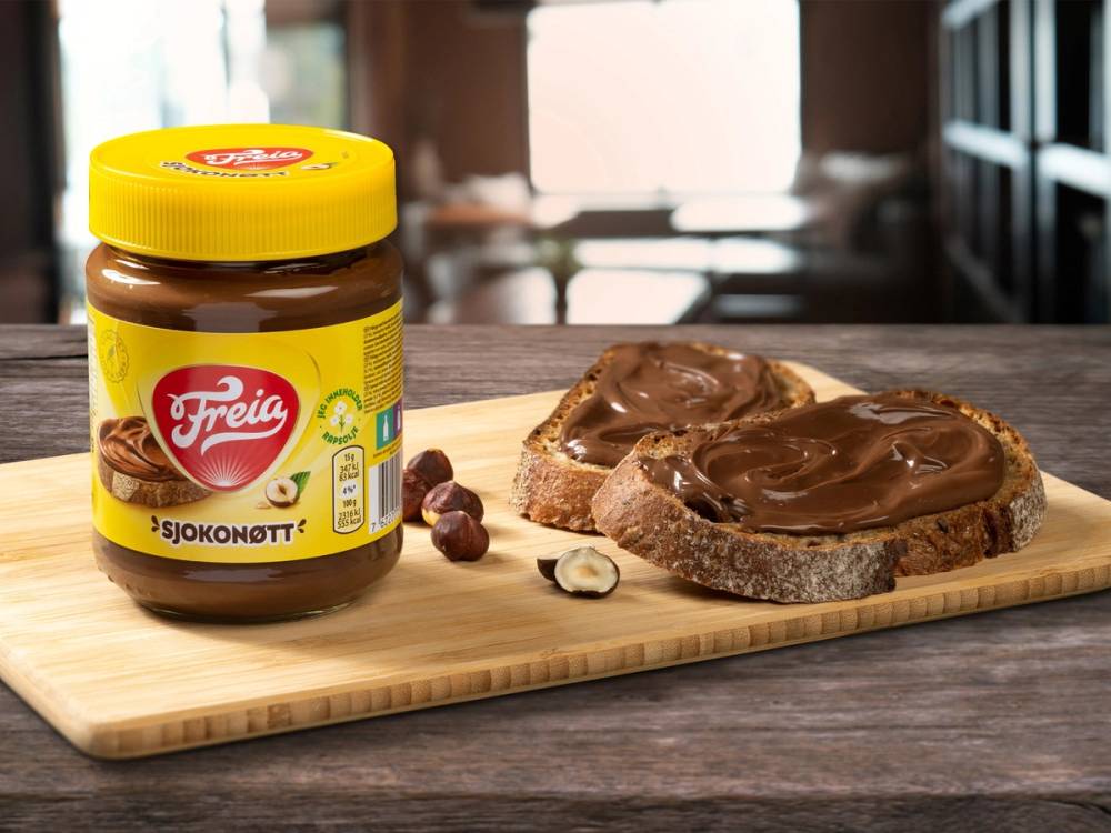 Et glass med sjokoladekrem fra Freia plassert ved siden av en skive brød med sjokoladekrem på en skjærebrett.