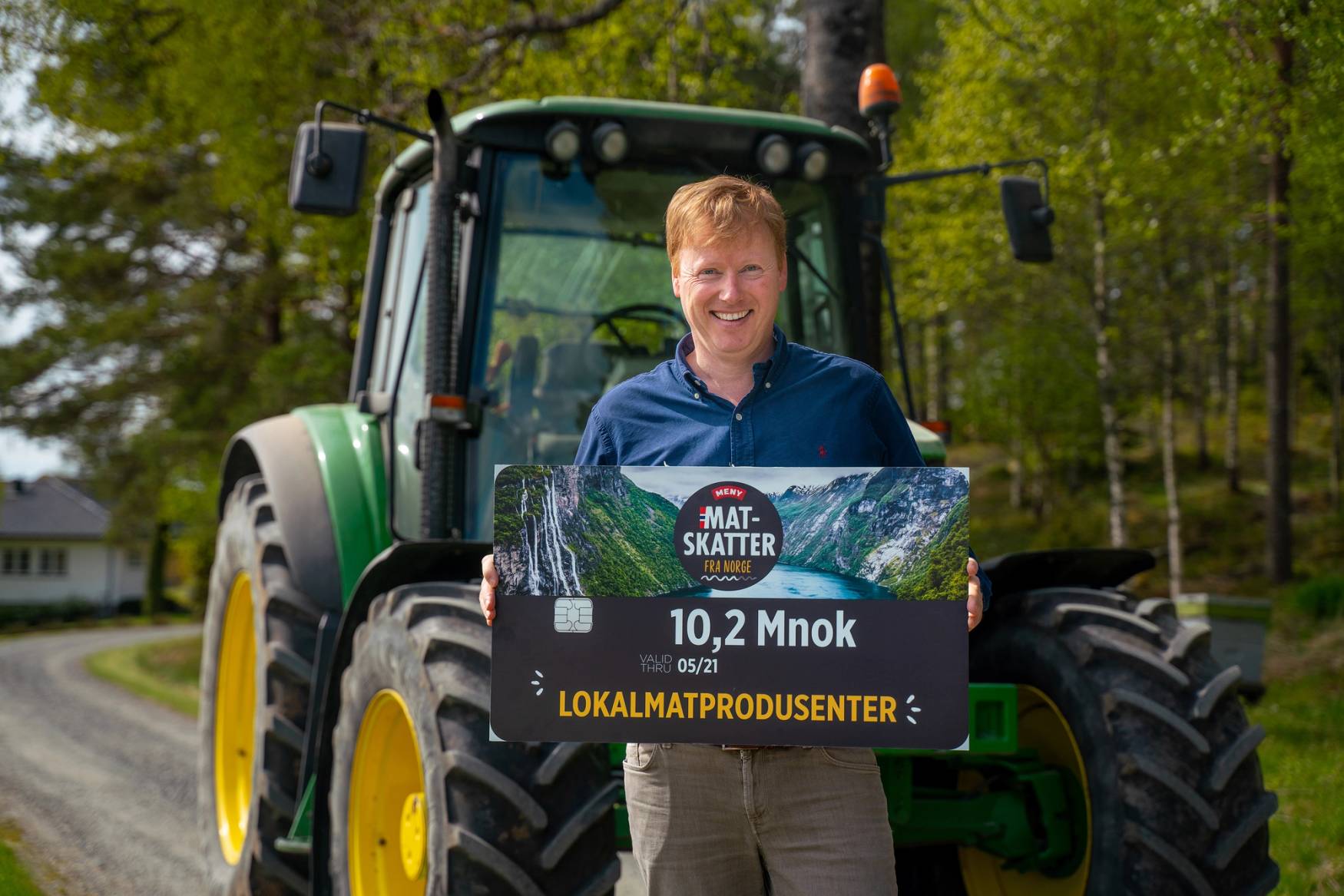 Person holder et stort sjekkhefte foran en traktor i en grønn skogkledd område, med teksten 'MAT SKATTER' og beløpet 10,2 millioner kroner.