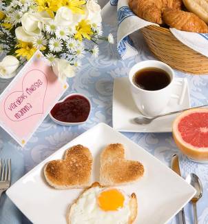 Hjerteformede toast og et speilegg på en hvit tallerken, med kaffe, frukt og syltetøy på et frokostbord.