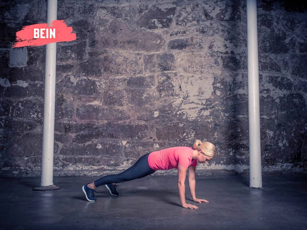 En kvinne i rosa treningstøy gjør burpees på gulvet i et treningsrom med en murvegg i bakgrunnen.