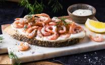 Rekesmørbrød med kremost, reker og dill på en skjærebrett, med sitron og en liten skål med saus ved siden av.
