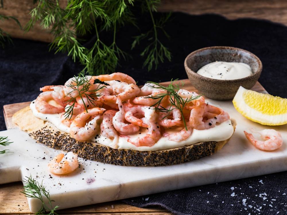 Rekesmørbrød med kremost, reker og dill på en skjærebrett, med sitron og en liten skål med saus ved siden av.