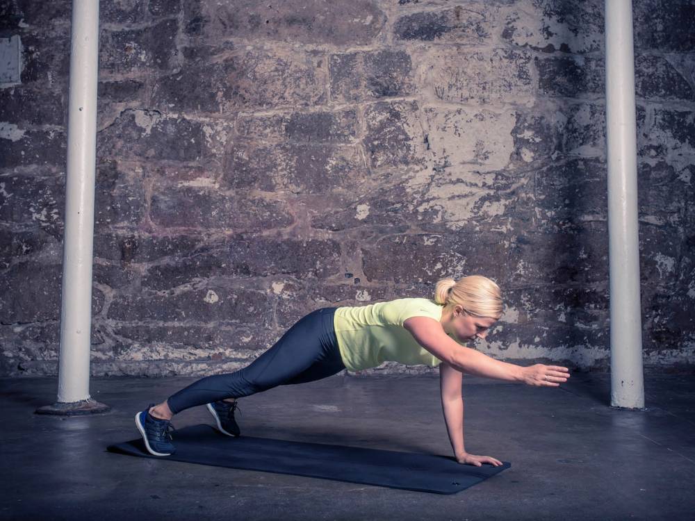 En person gjør pushups med vridning på en treningsmatte i et treningsrom med murvegg.