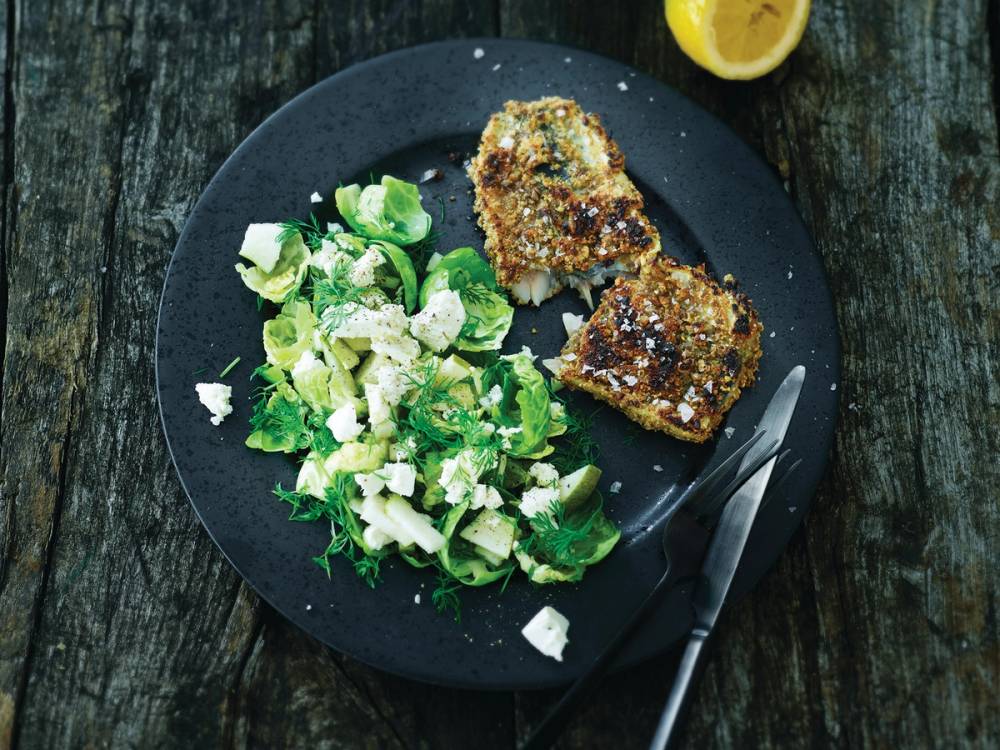 To brødskiver med pålegg og en grønn salat på en svart tallerken, med en sitronhalvdel ved siden av, på et rustikt trebord.