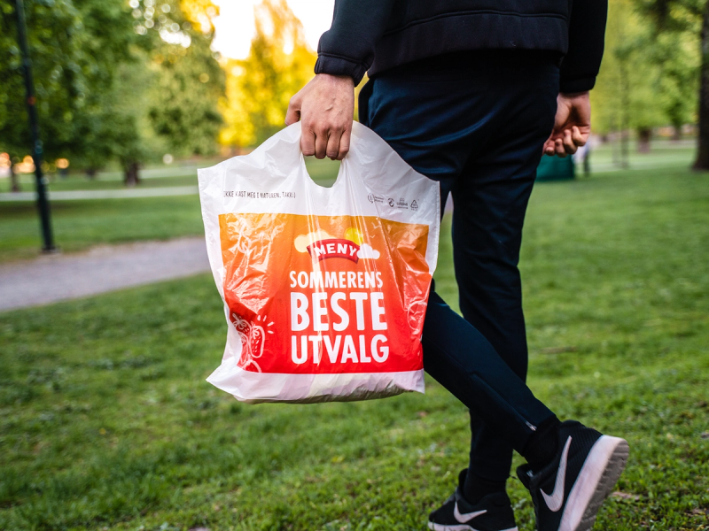 Person som går i en park med en plastpose med teksten 'SOMMERENS BESTE UTVALG' i hånden.