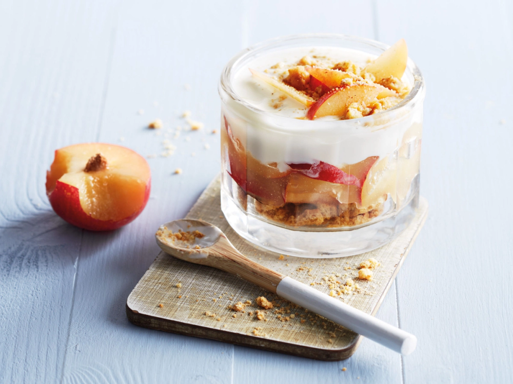 En glass med havrekjeks og plommer, ser ut til å være en dessert eller fruktkompott, plassert på et lite skjærebrett med en skje ved siden av.