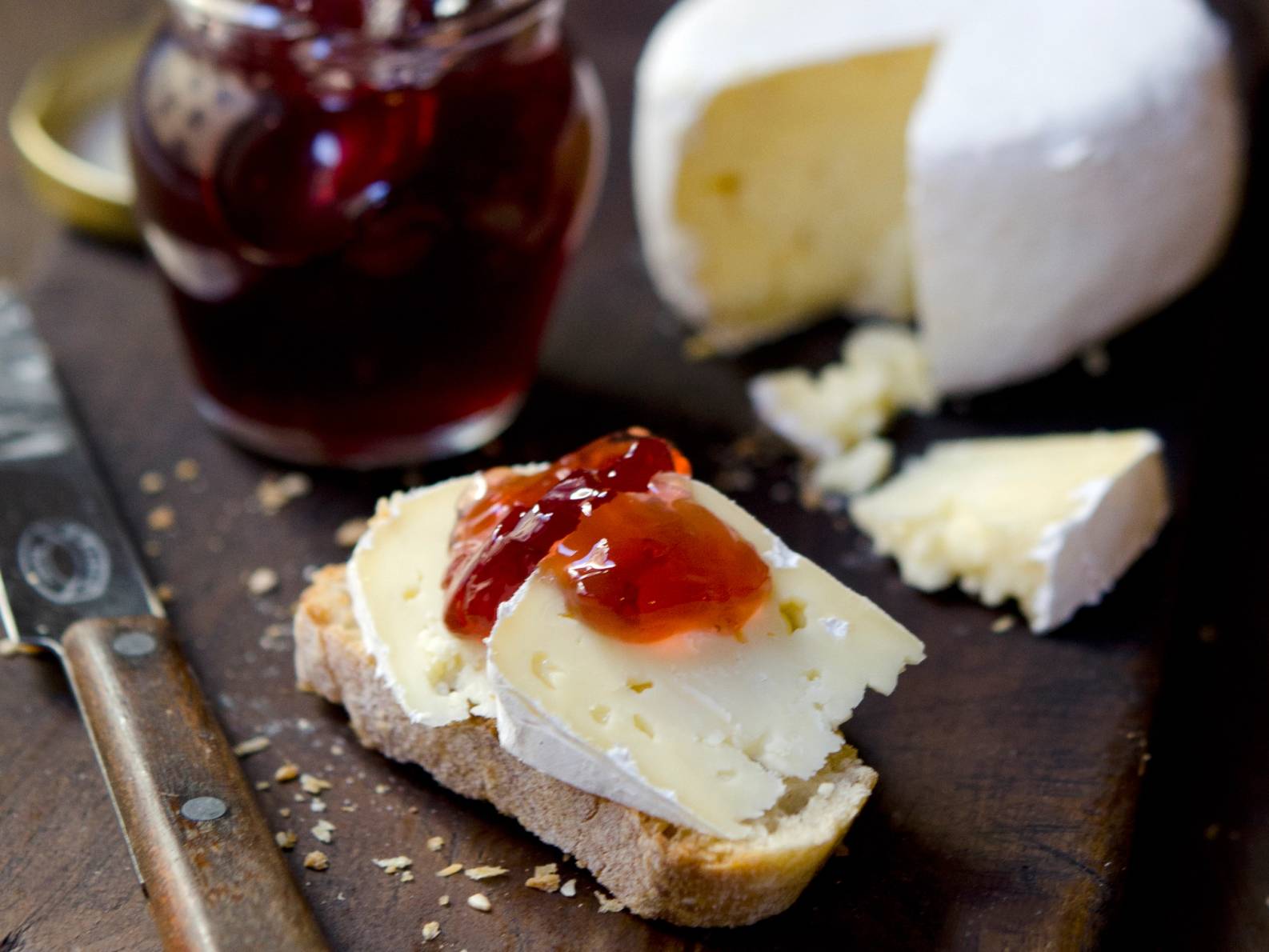 Et stykke brød med smør og jordbærsyltetøy, omgitt av en ost og en skje, på et treplate med en glass med rød drikke i bakgrunnen.