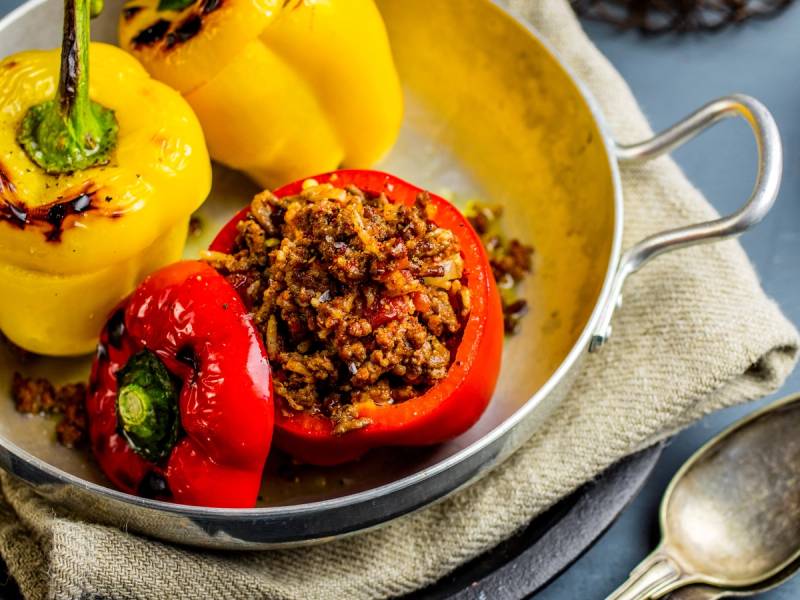 Fylte paprikaer med ris og kjøttdeig i en stekepanne, omgitt av gule paprikaer og en skje på siden.