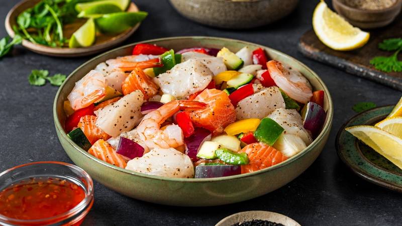En bolle med en salat bestående av scampi, agurk, paprika og rødløk, omgitt av små skåler med lime, krydder og saus.