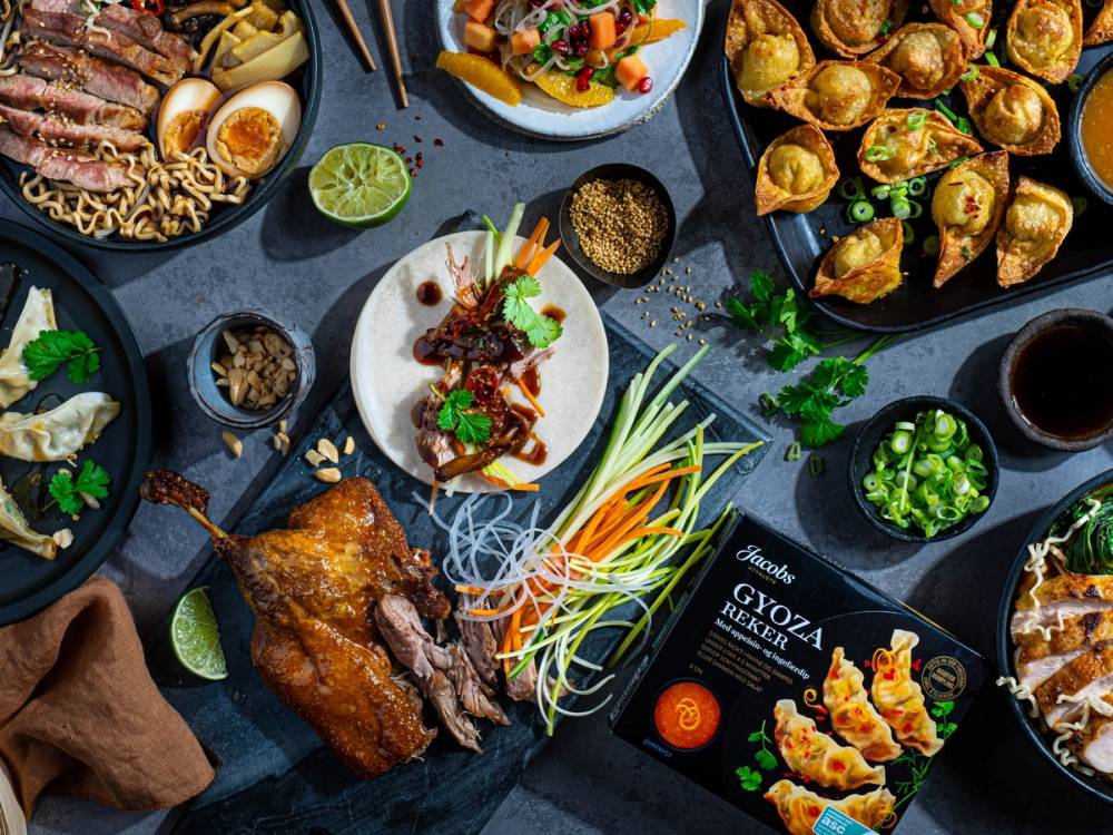 Et utvalg av asiatiske retter og snacks på en svart bordplate, inkludert vårruller, kylling, grønnsaker og dip, med noen limebåter og urter.