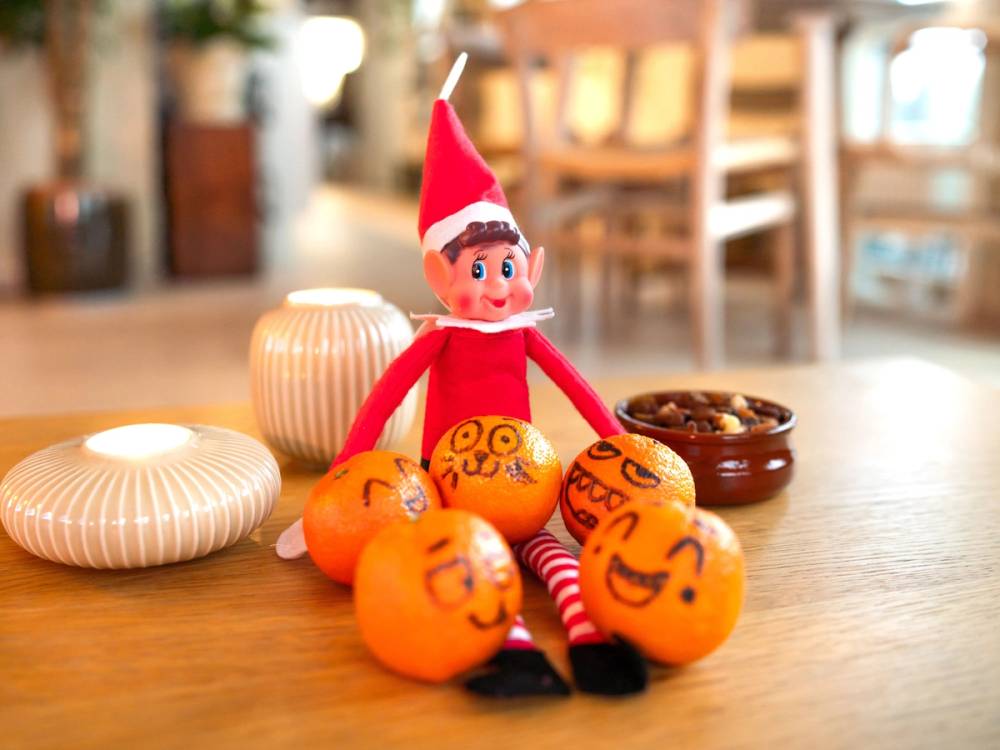 En liten nissefigur med rød lue og klær omgitt av juledekorasjoner og små gule klementiner med ansikter på et bord.