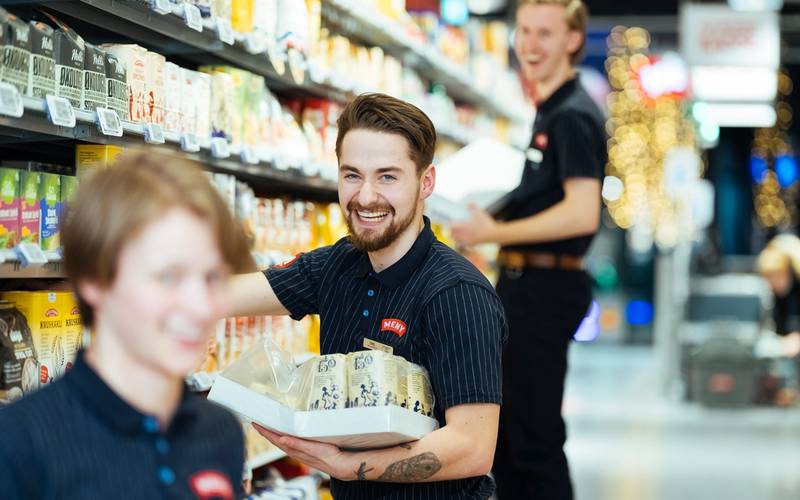 En person med svart skjorte handler i en dagligvarebutikk med hyller fylt av produkter.