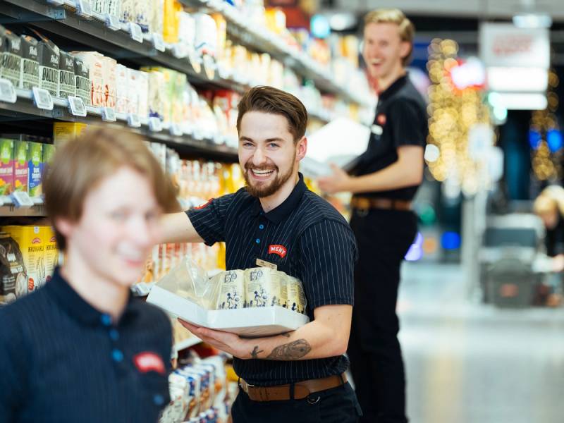 En person med svart skjorte handler i en dagligvarebutikk med hyller fylt av produkter.