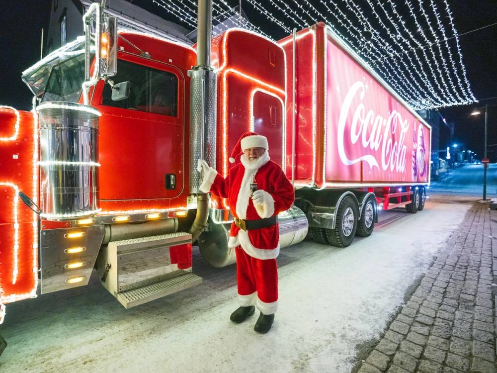 En person kledd som julenisse står foran en stor, pyntet Coca-Cola-lastebil med julelys på en vinterkveld.