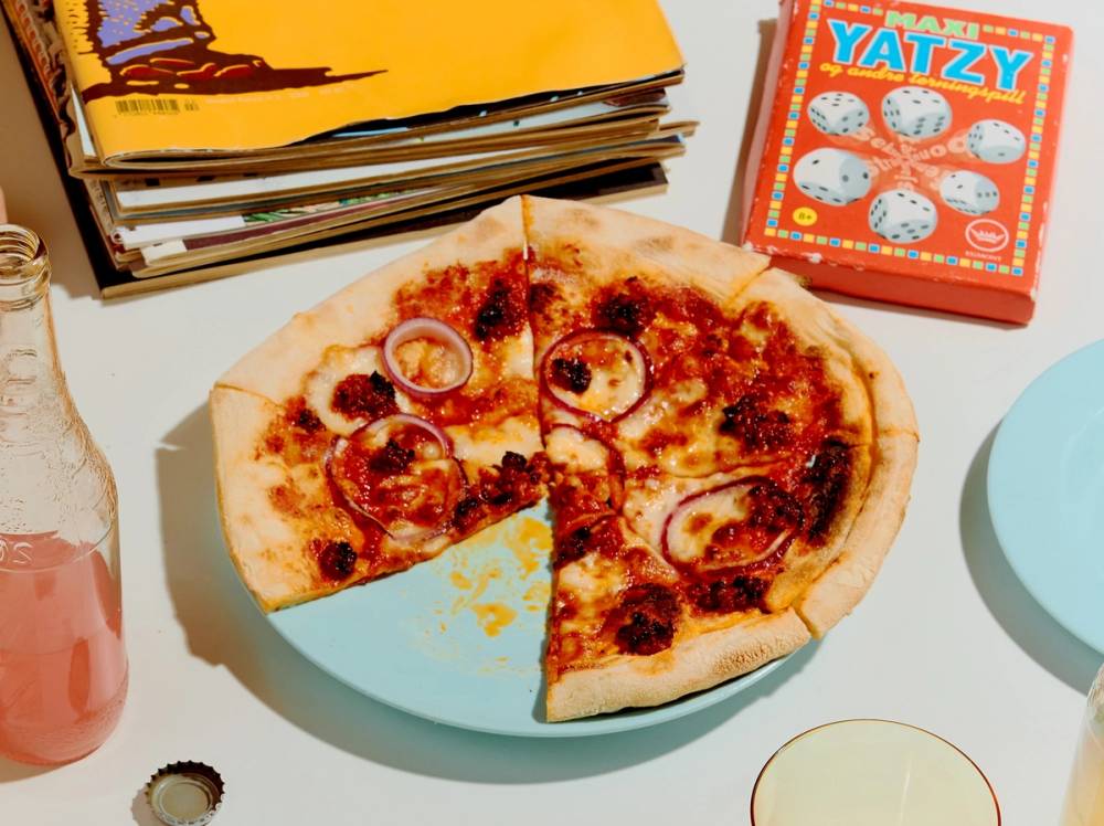 En pizza med ost og skinke på et bord med ulike spill og bøker rundt.