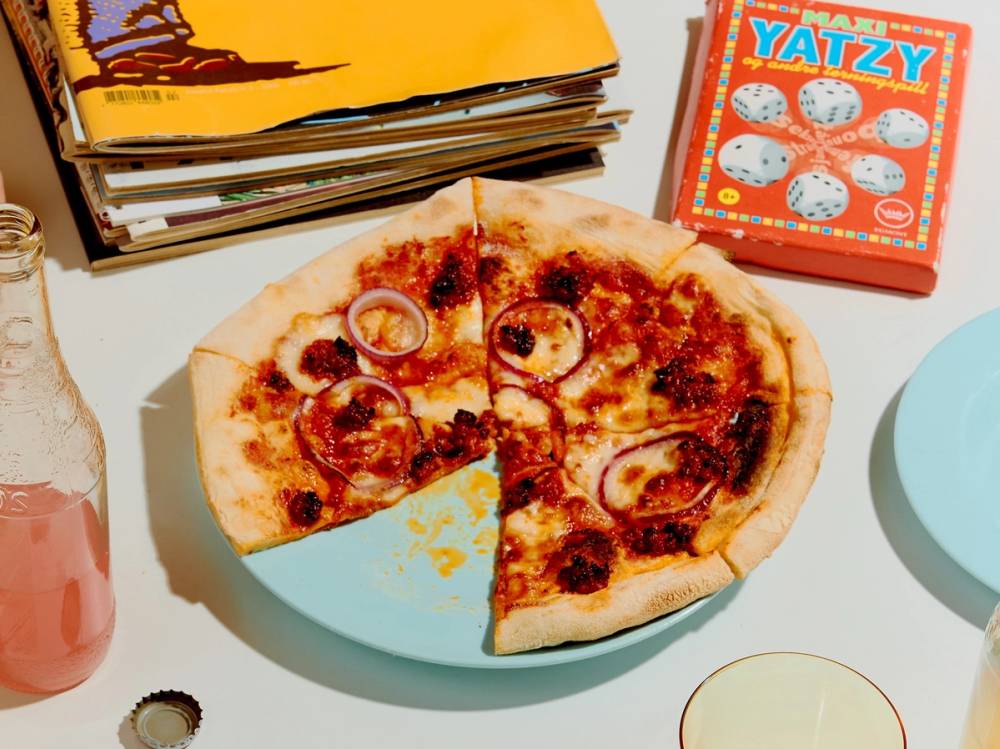 En pizza med ost og skinke på et bord med ulike spill og bøker rundt.