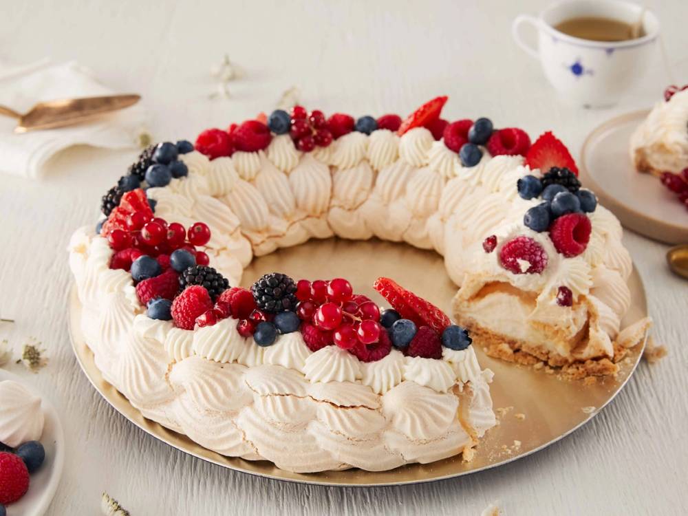 En pavlova krans pyntet med blåbær, bringebær og krem, med et stykke tatt ut av den på en tallerken.