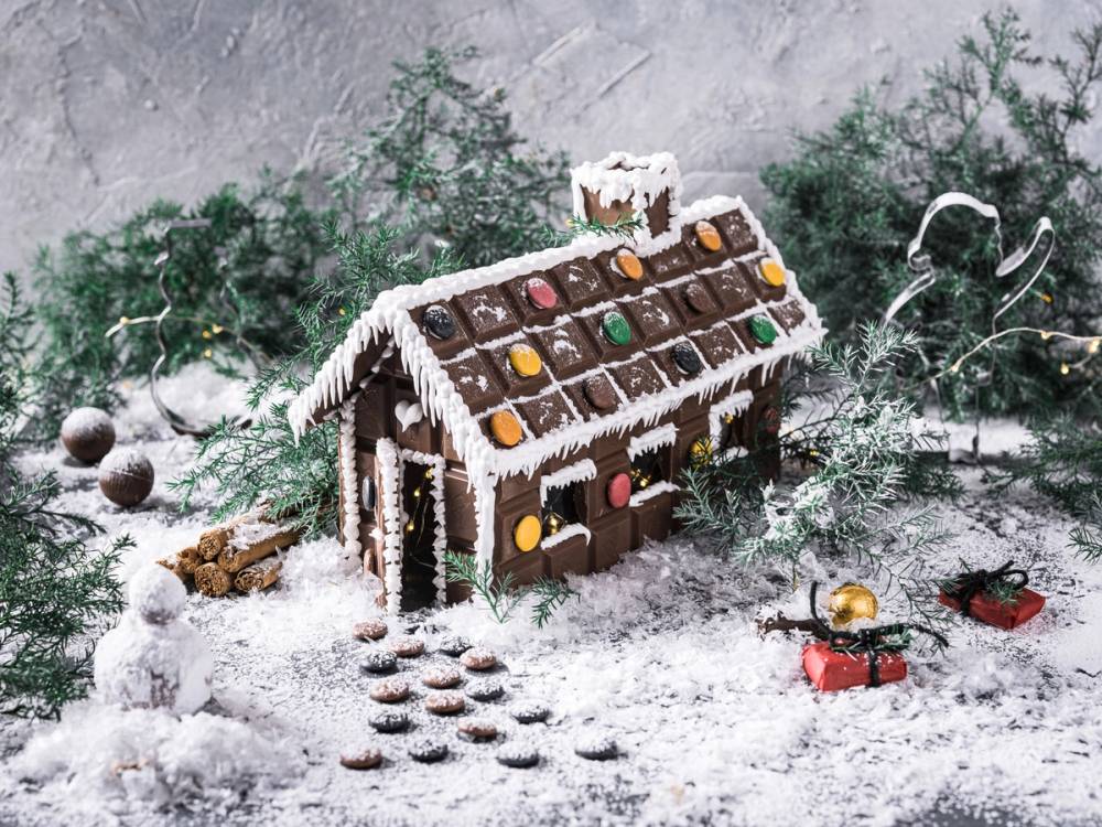 Et lite hus laget av sjokolade med pynt og snø rundt, omgitt av grantrær og julepynt.