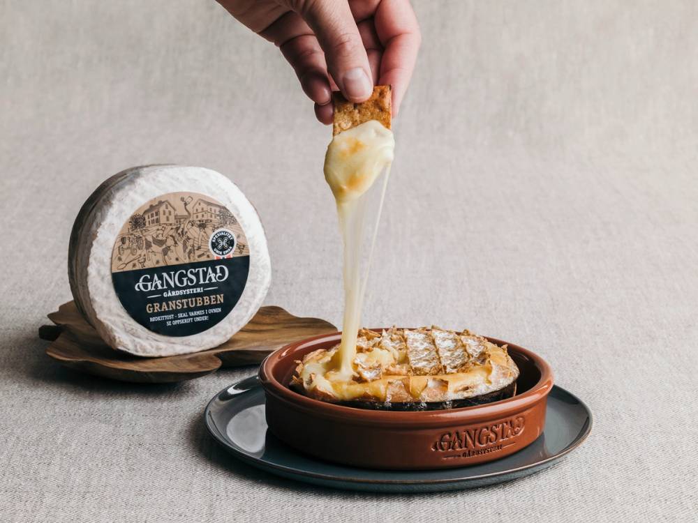 En person dypper en ostestav i en gryte med gratinert mat, med en osteklokke på bordet ved siden av.