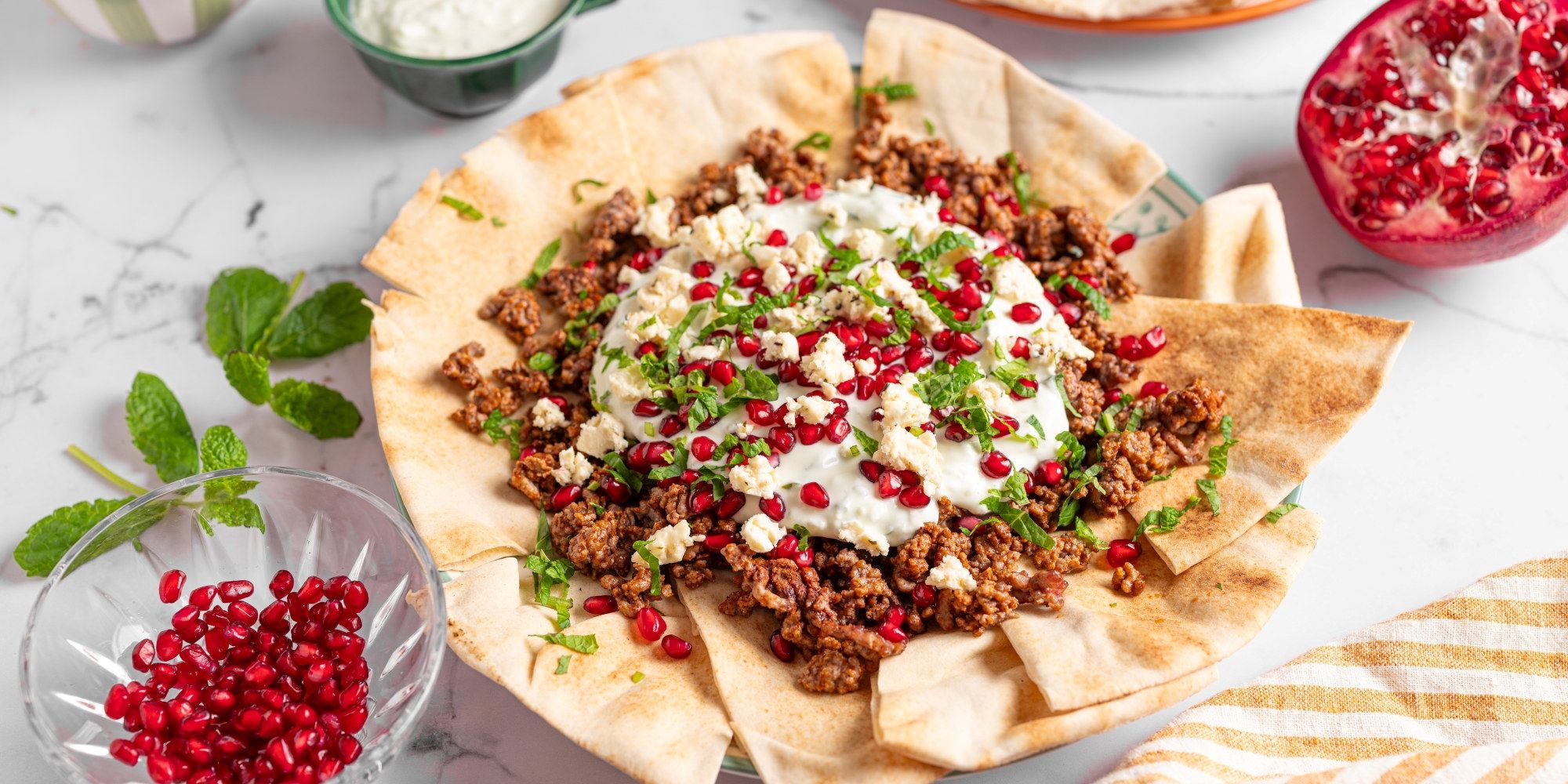 Liba brød med lamme kjøttdeig, toppet med yoghurt, granateple og frisk mynte, på et hvitt bord.