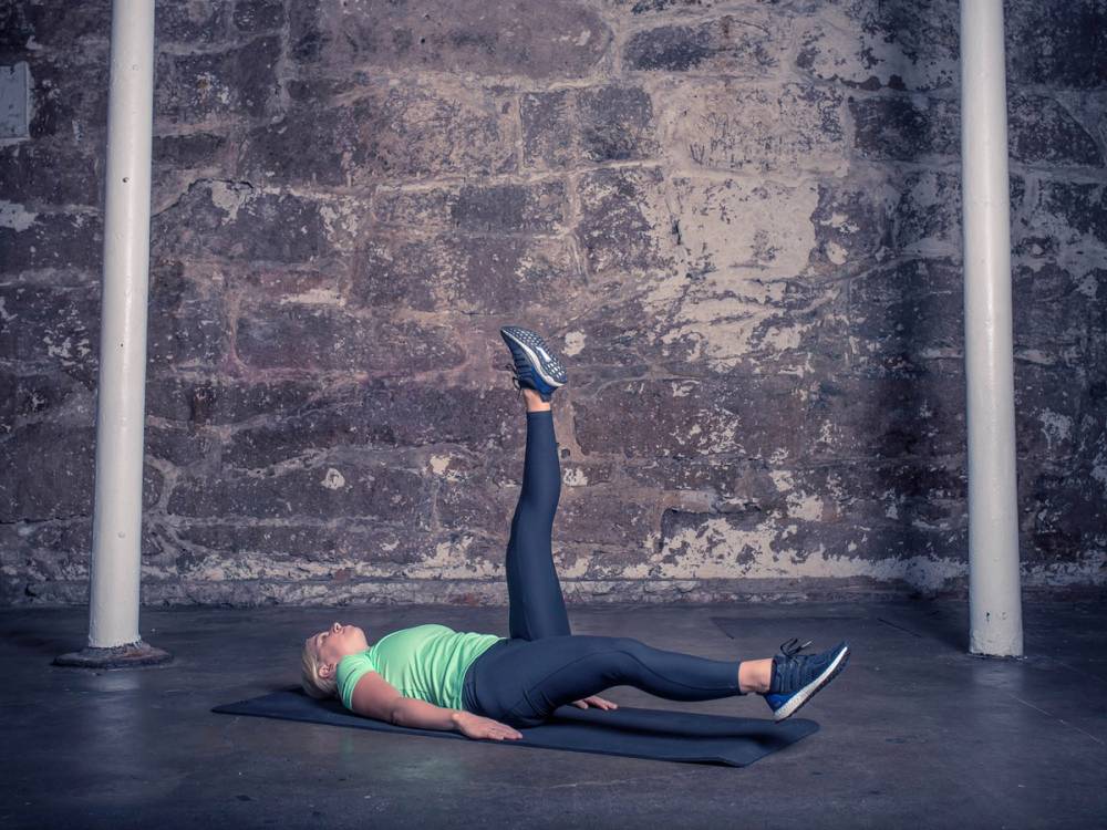 En kvinne ligger på ryggen på en treningsmatte og strekker ut bena i en øvelse mot en murvegg.