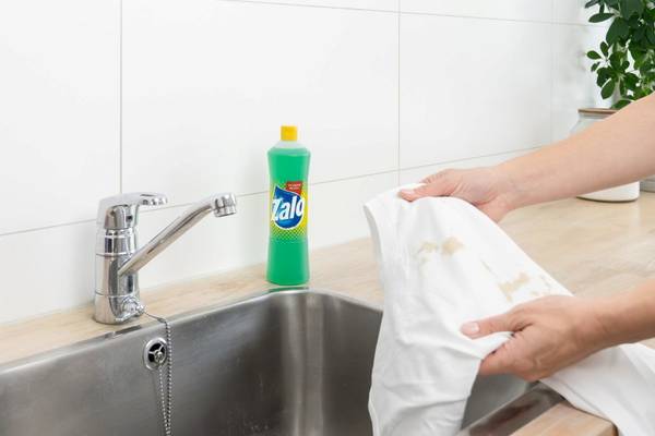 Person som bruker flekkfjerner på en hvit klut over kjøkkenvasken, med en flaske Zalo flytende oppvaskmiddel på benken.
