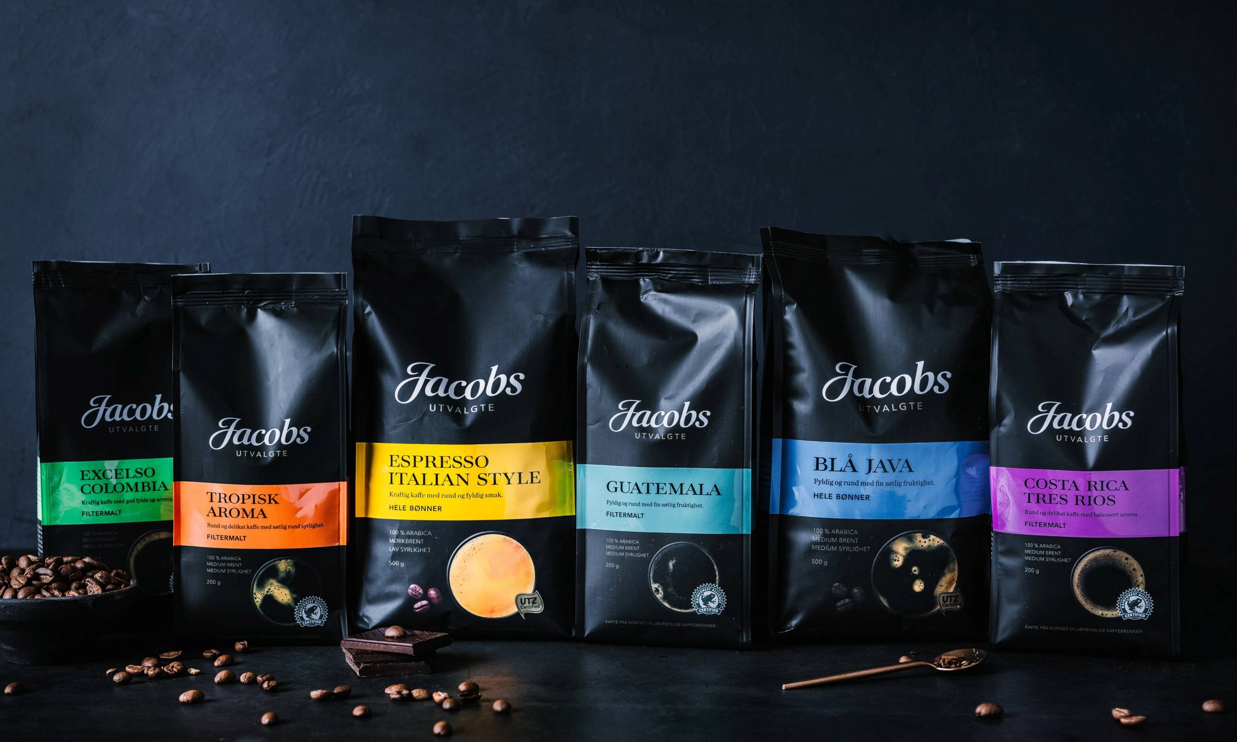 Ulike typer kaffe i svarte poser med fargerike etiketter, plassert på en mørk bakgrunn med noen kaffebønner rundt.