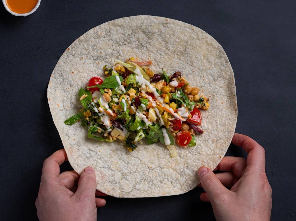 En person holder en tortilla med salat og grønnsaker, klar til å lage en wrap, på en mørk bakgrunn.