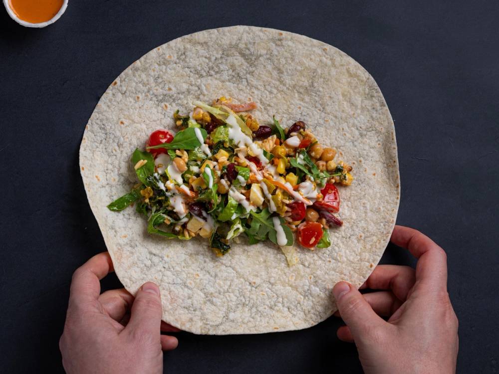 En person holder en tortilla med salat og grønnsaker, klar til å lage en wrap, på en mørk bakgrunn.